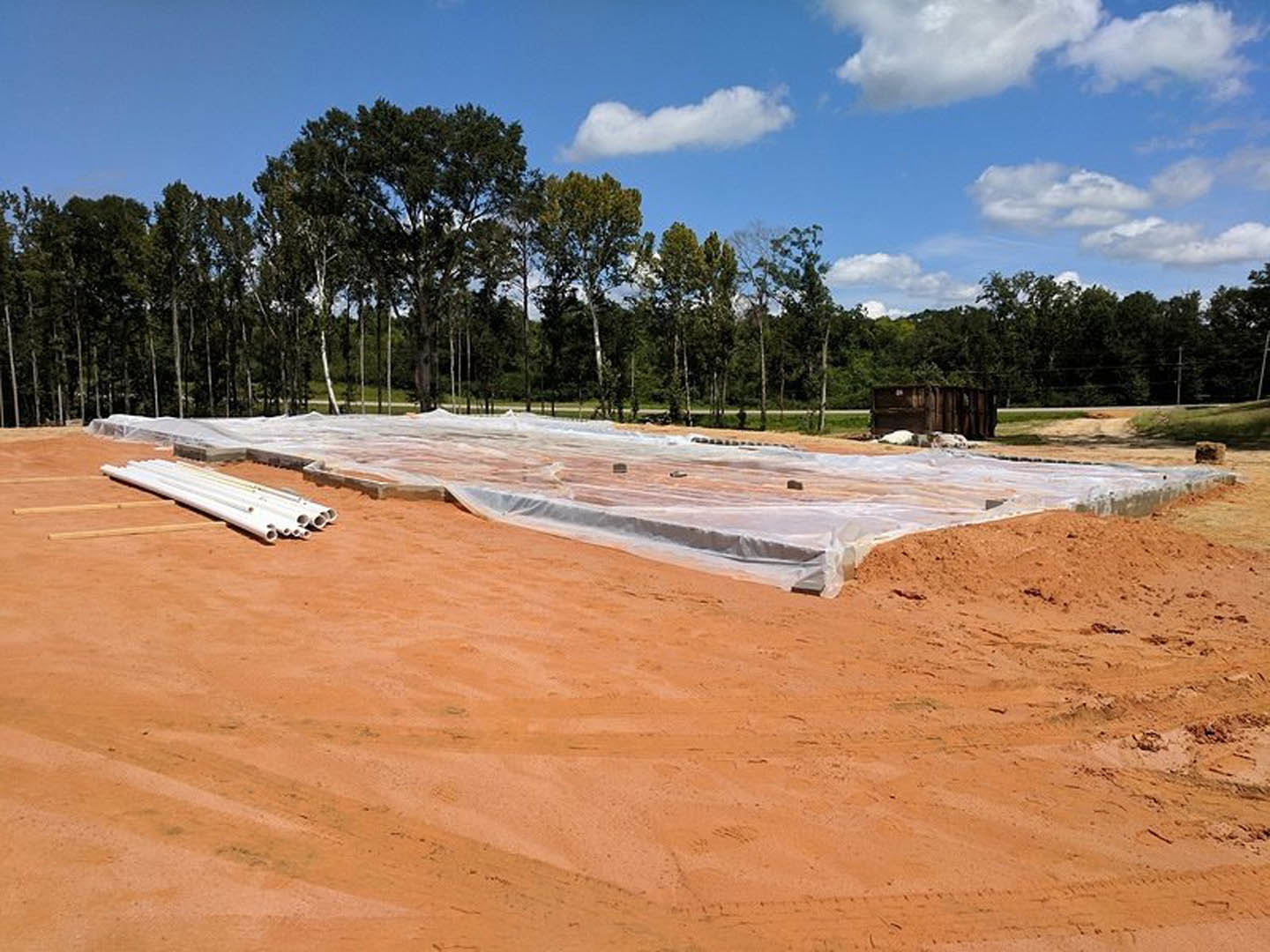 Plastic tarp covering sandy soil at a residential construction site, with exposed white pipes and a partially built wall in the background under a cloudy sky.