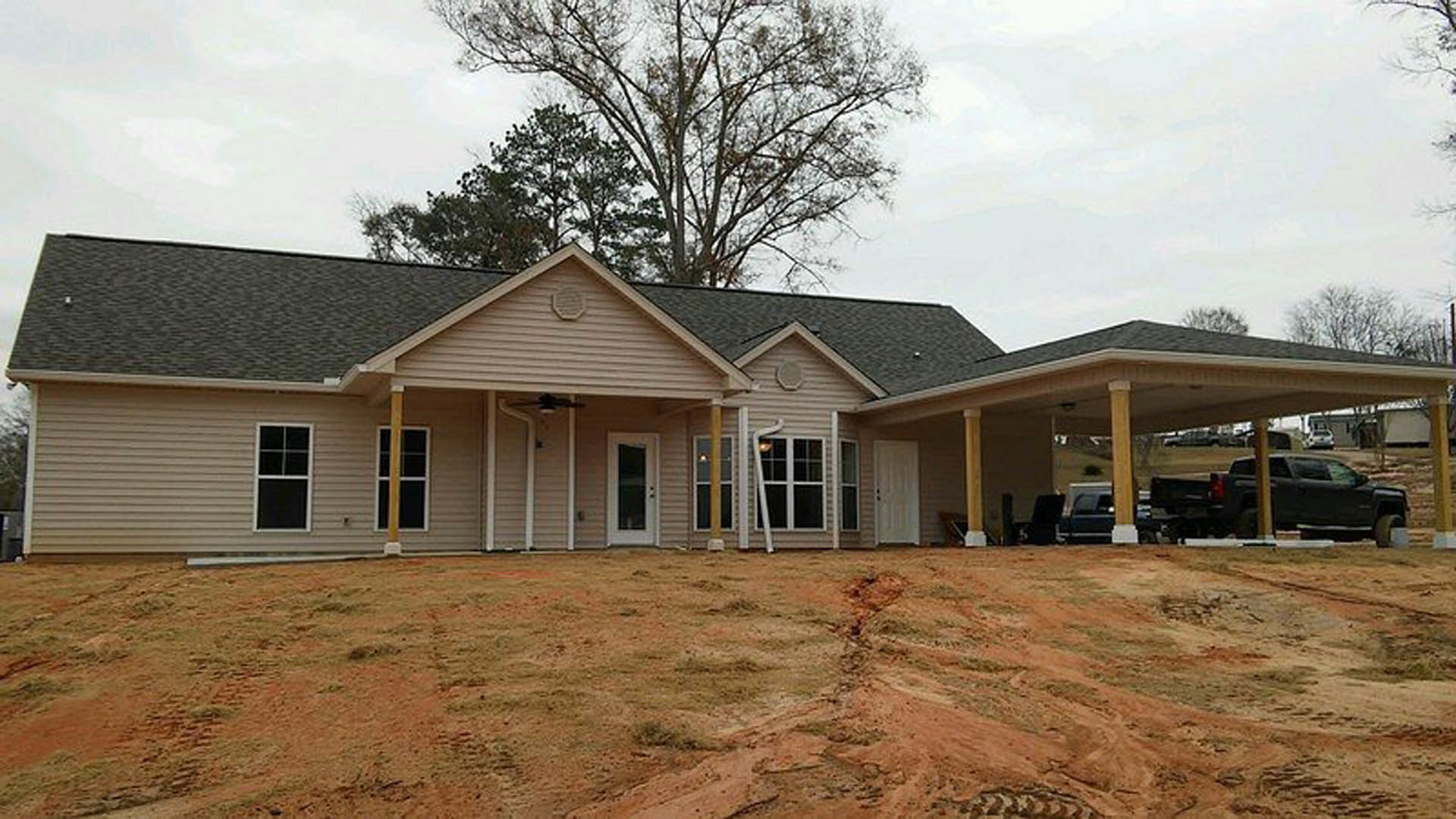 Two-story home with gray siding, covered front porch, white trim, and large windows, set on a dirt lot with a white fence and mature tree