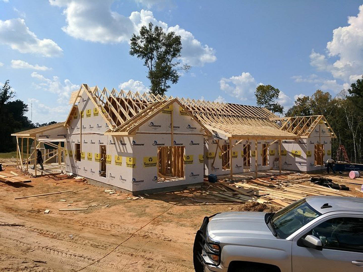 Wood-framed house under construction with pickup truck parked in front, leafy tree nearby, person working on site, cloudy sky overhead