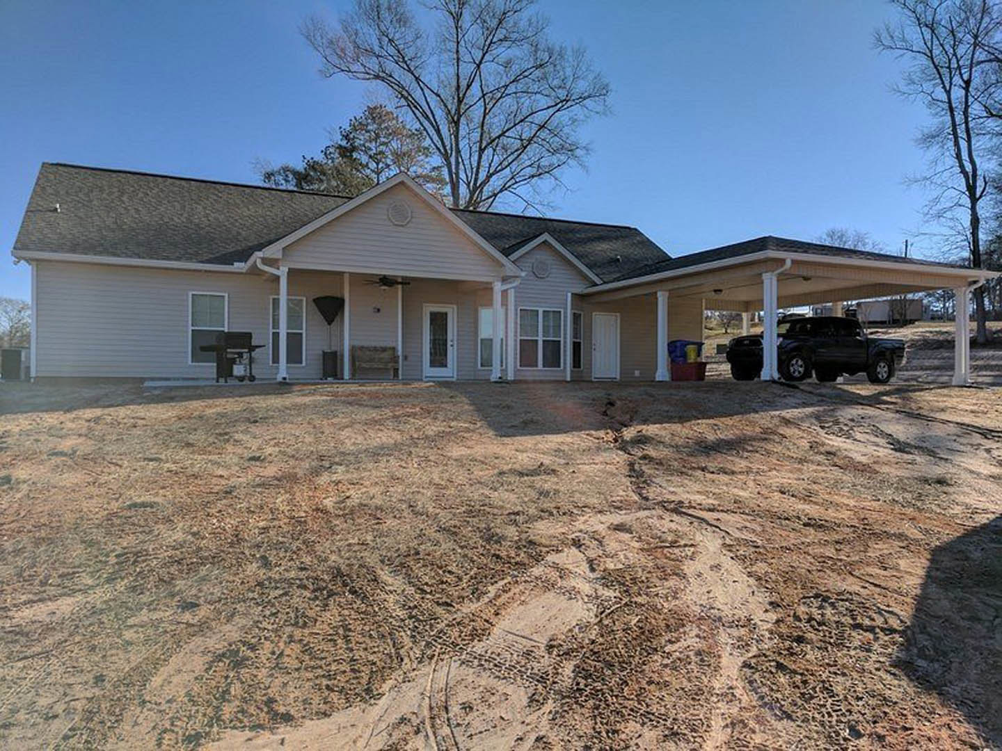 Wide concrete driveway leading to a modern house with a patch of bare dirt, leafless tree, black car parked near a white pole, and front door featuring a window.