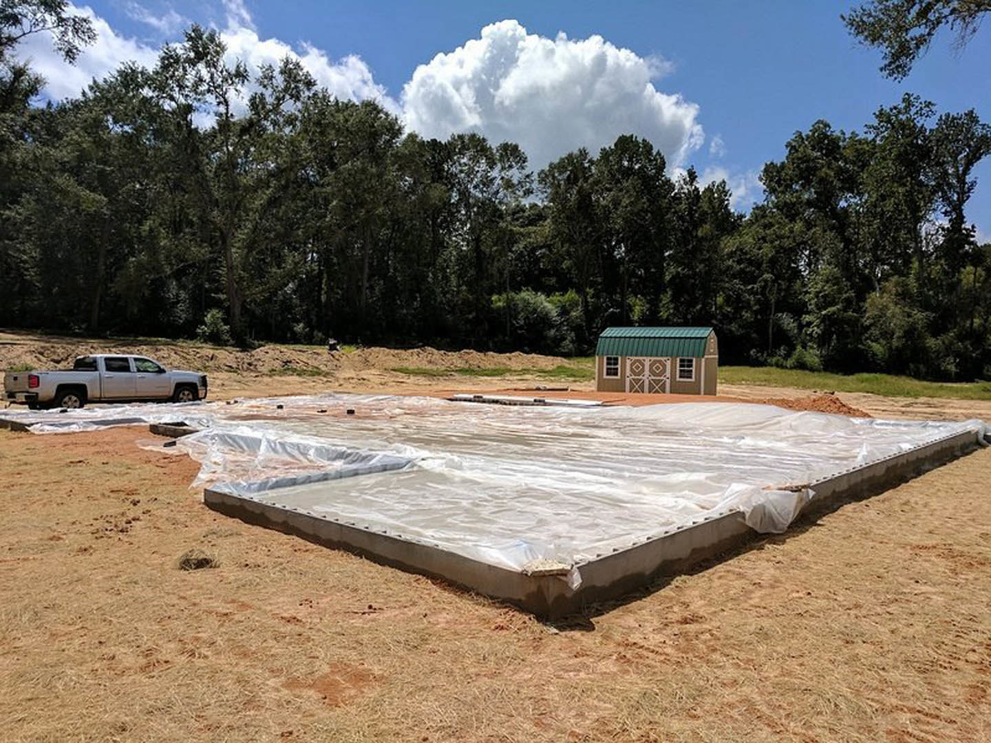 Dirt construction site with green-roofed building, white truck parked near tarp-covered foundation, trees and clouds in background