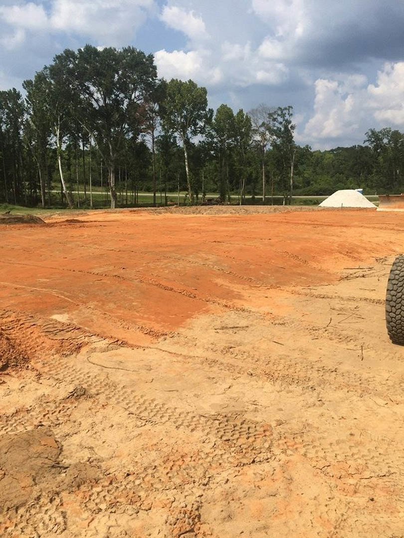 Expansive dirt field with visible tire tracks, bordered by leafy trees under a clear blue sky