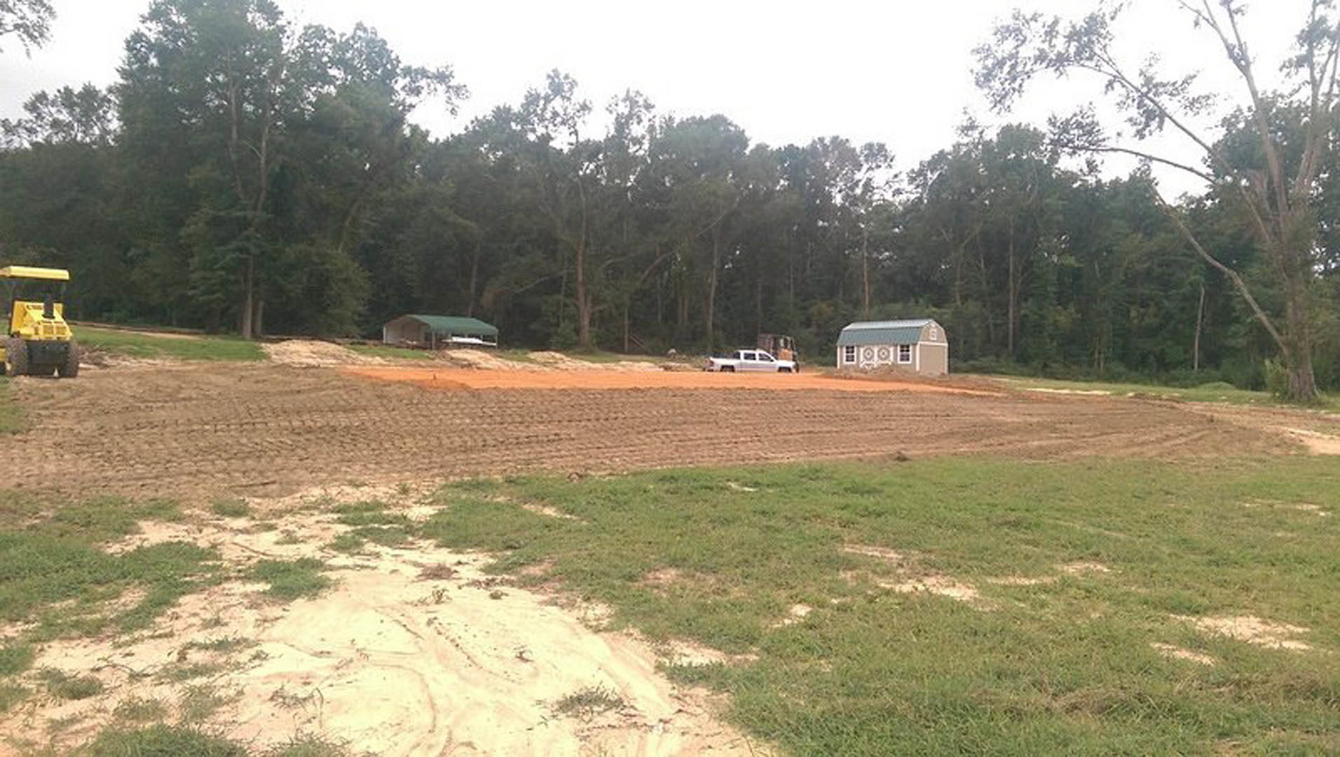 Modern home with green roof surrounded by grassy field, mature trees in background, yellow tractor and pickup truck parked nearby on dirt and grass.