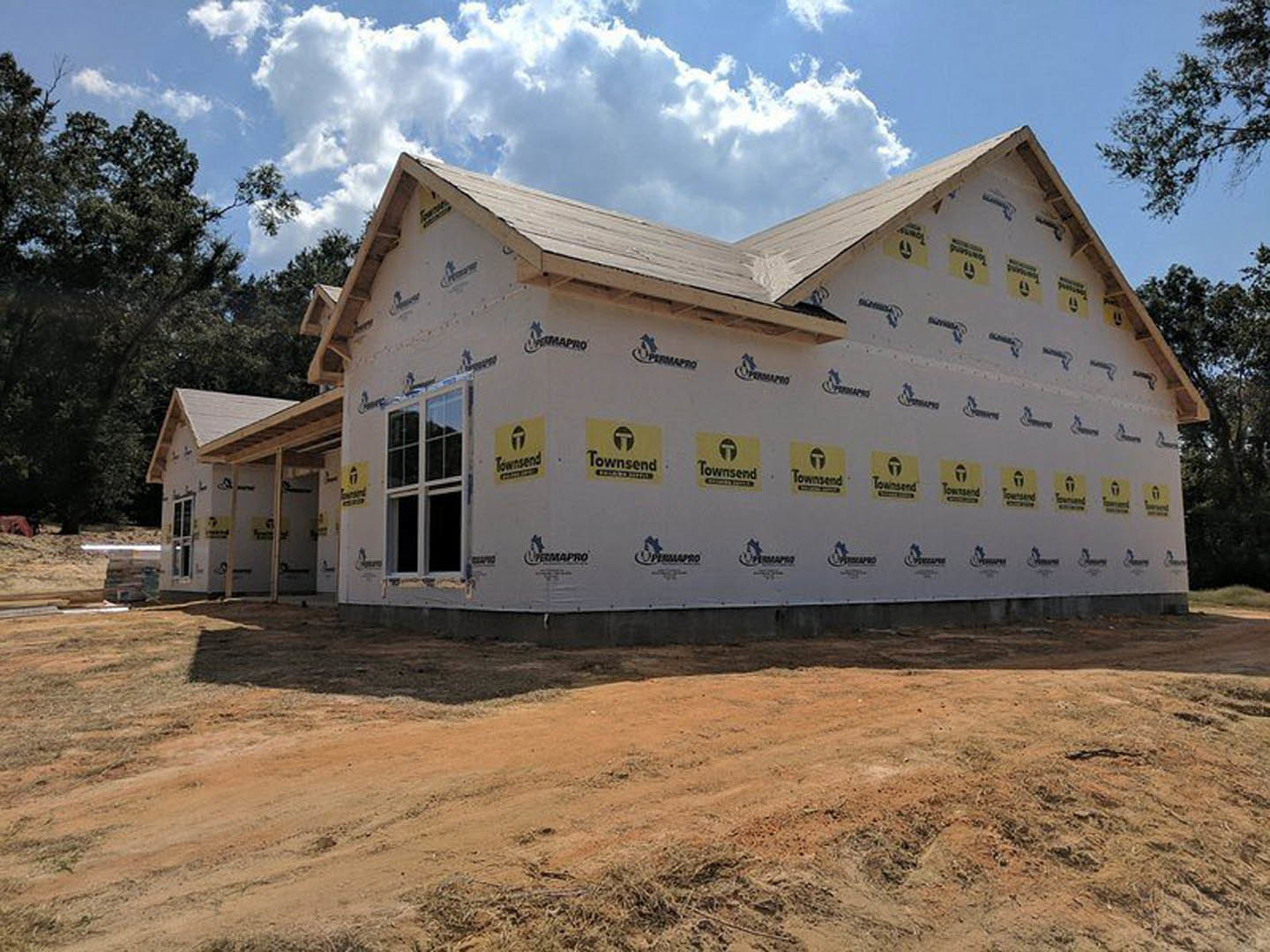 Two-story house under construction with exposed framing, white siding partially installed, construction signs attached to exterior wall, dirt driveway in foreground, leafy tree