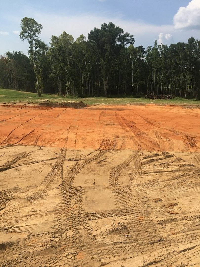 Dirt field with tire tracks bordered by trees under a partly cloudy sky