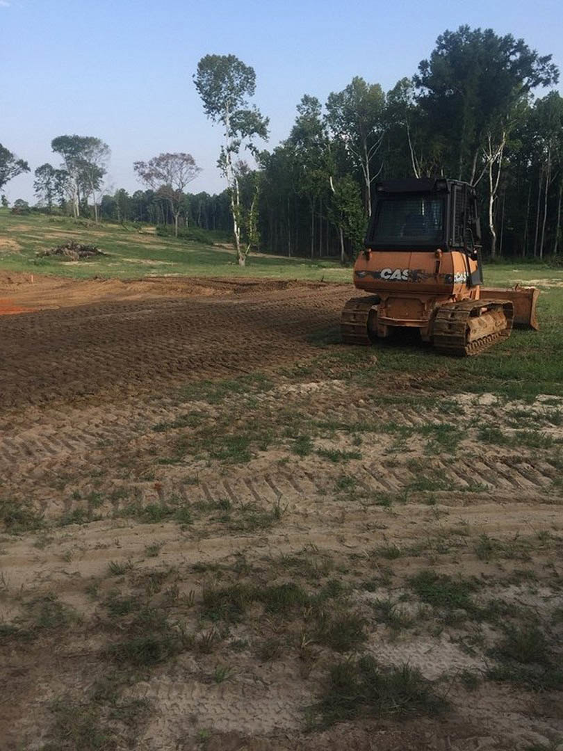 Modern farmhouse with white siding and black-framed windows, surrounded by grassy field and mature trees, blue sky overhead, tractor parked on soil near the home.