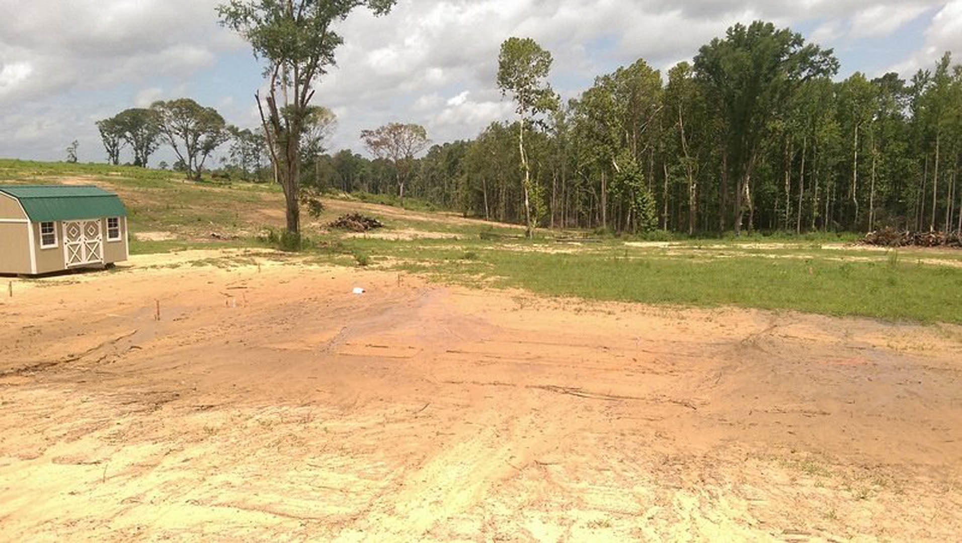 Dirt field bordered by trees, small shed with green roof, patches of grass, white object near center, dirt road leading toward forested background under cloudy sky