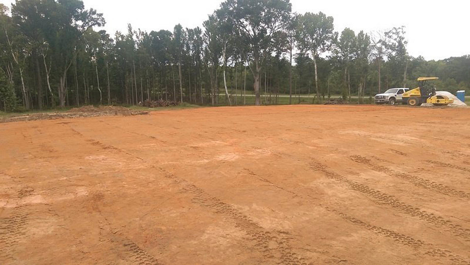Dirt field with tire tracks bordered by a group of trees, yellow construction vehicle with large wheel and cab, white truck with person in window, clear sky above
