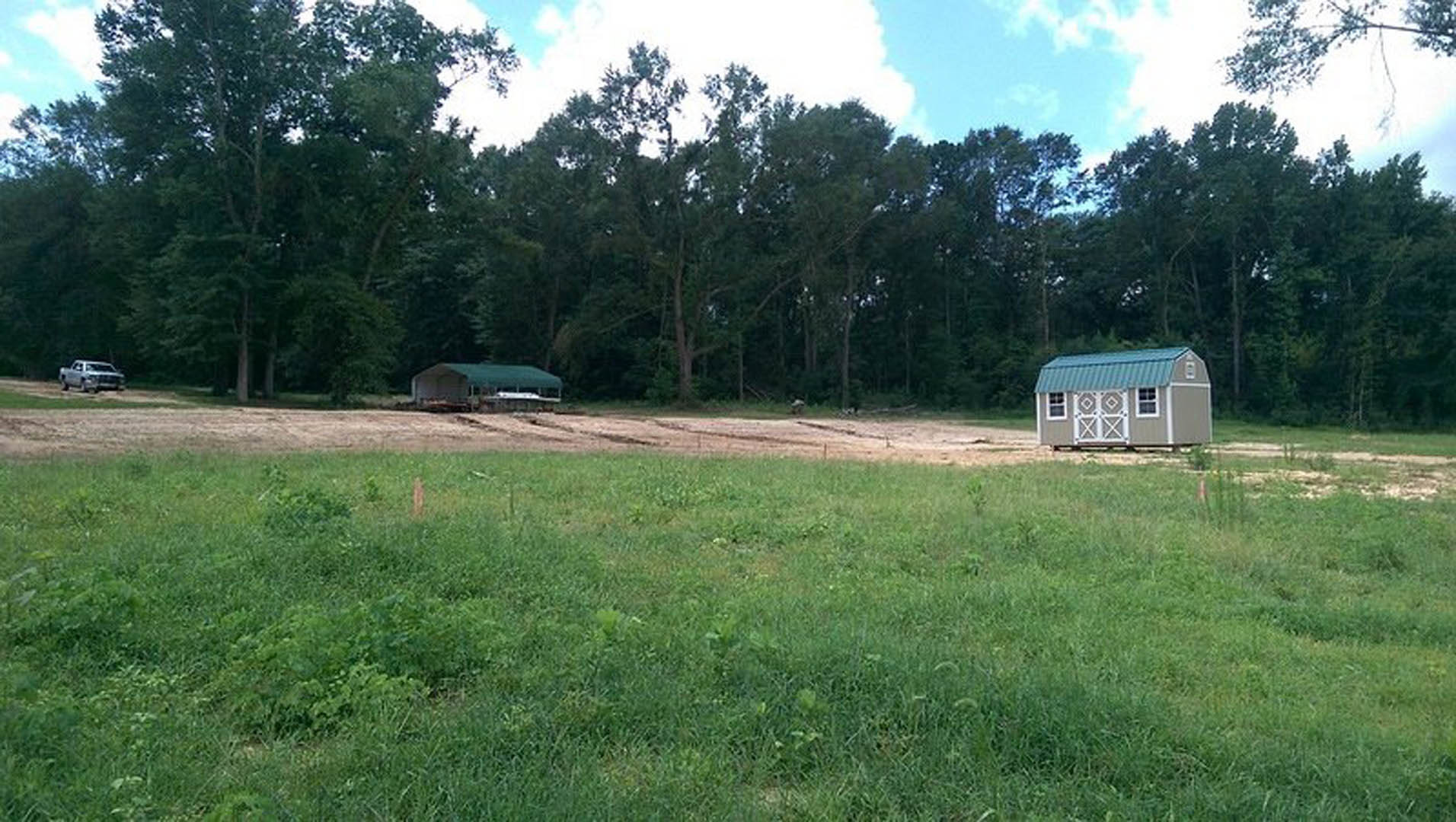Small blue-roofed shed on a grassy field with scattered dirt patches, silver truck partially visible, dense cluster of trees in the background under a cloudy sky