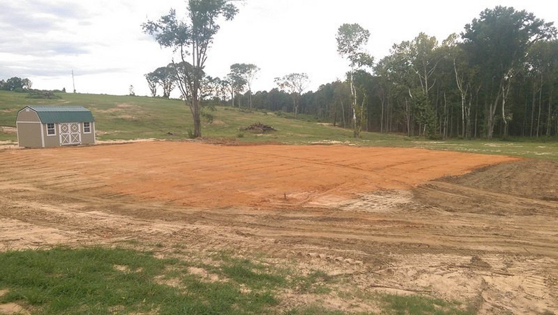 Dirt field bordered by trees, green-roofed building in the background, wooden fence along the edge, scattered grass patches, cloudy sky overhead
