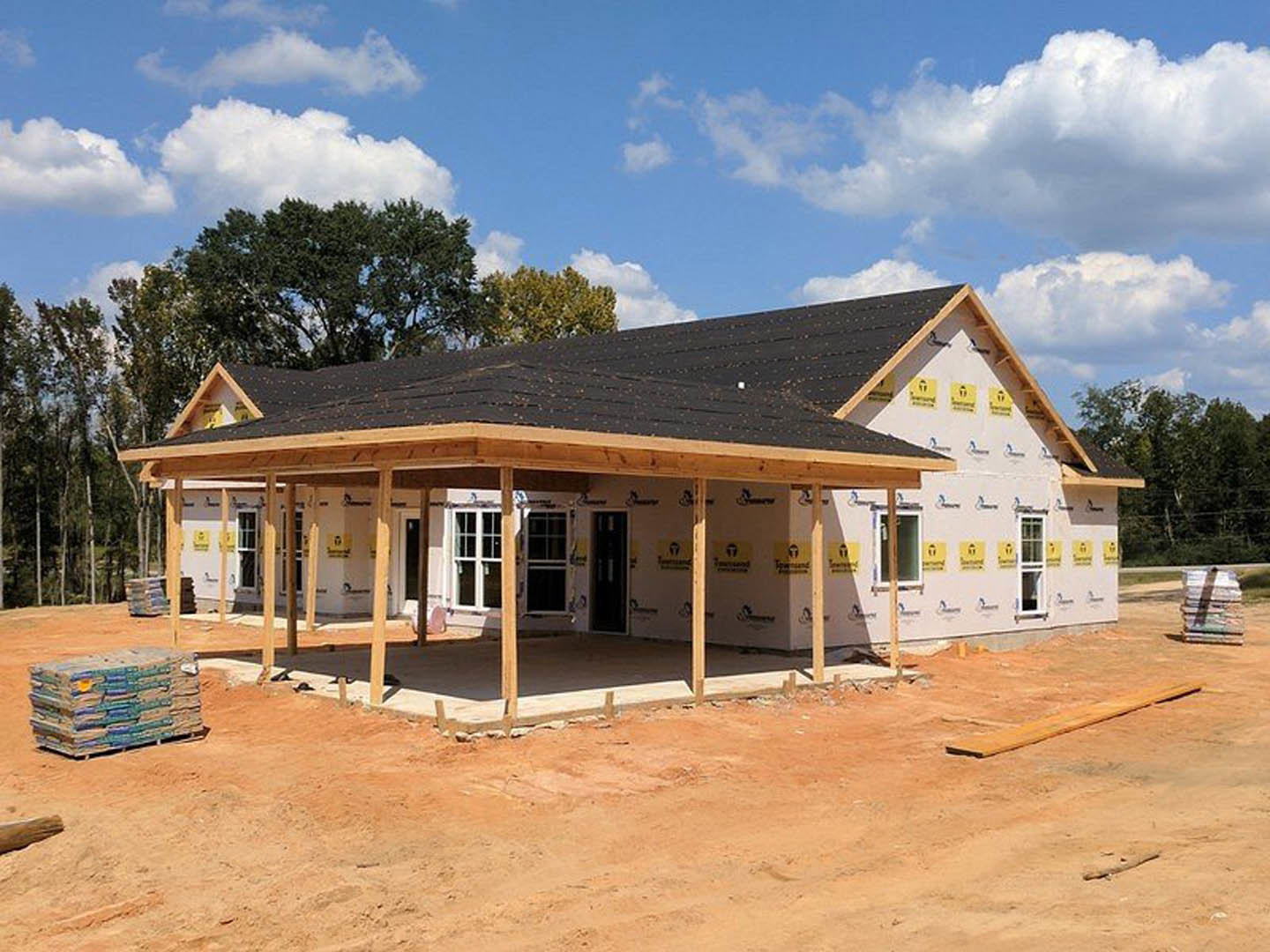 Two-story house under construction featuring a spacious covered porch, black shingle roof, white-framed windows, exposed framing, and dirt yard with construction materials stacked