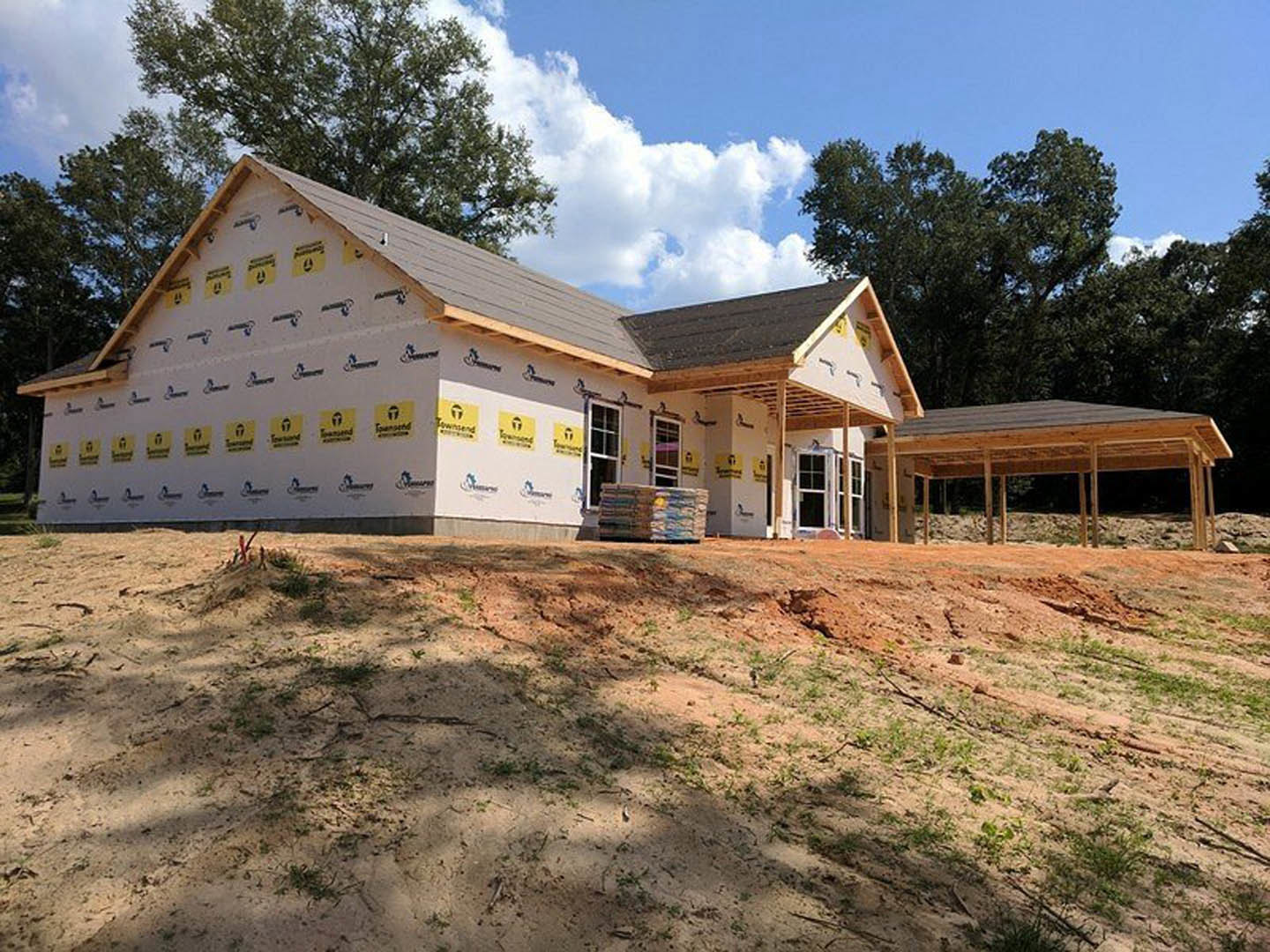 Wood-framed house under construction with covered porch, exposed beams, and unfinished exterior walls, surrounded by dirt lot and scattered construction materials.