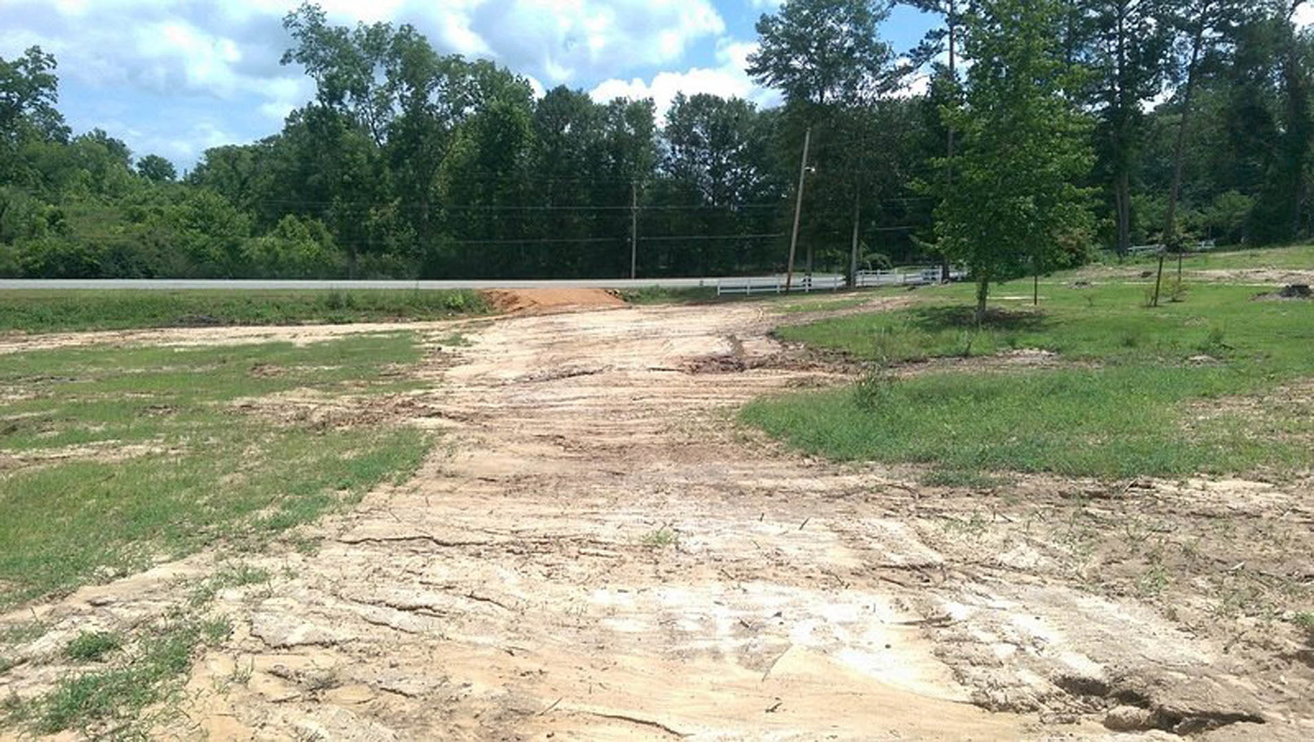 Dirt road bordered by grass and white fence, group of trees and power lines in background under cloudy sky