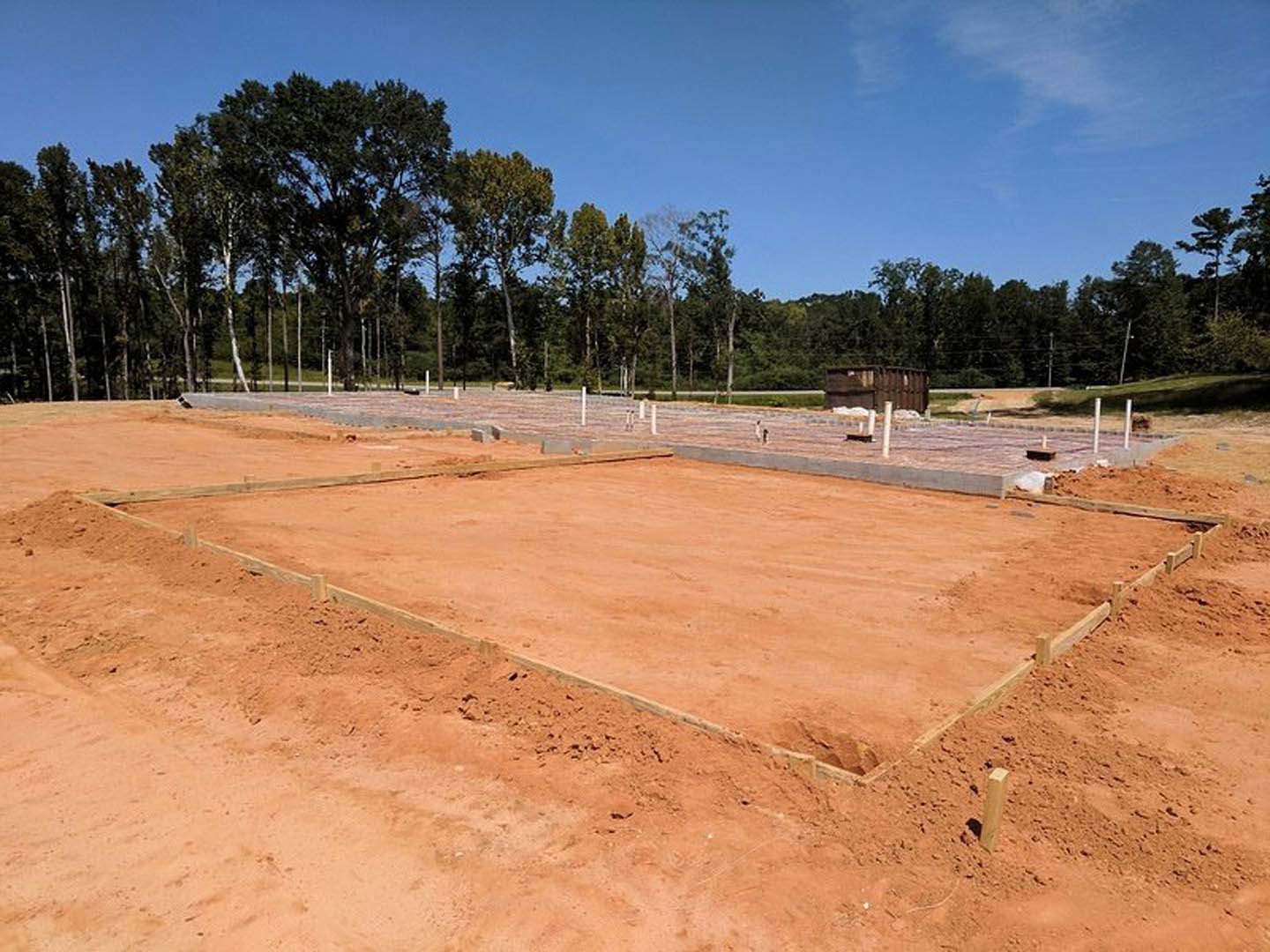 Concrete foundation surrounded by dirt and soil, construction materials scattered, trees and blue sky in background, temporary fencing along perimeter