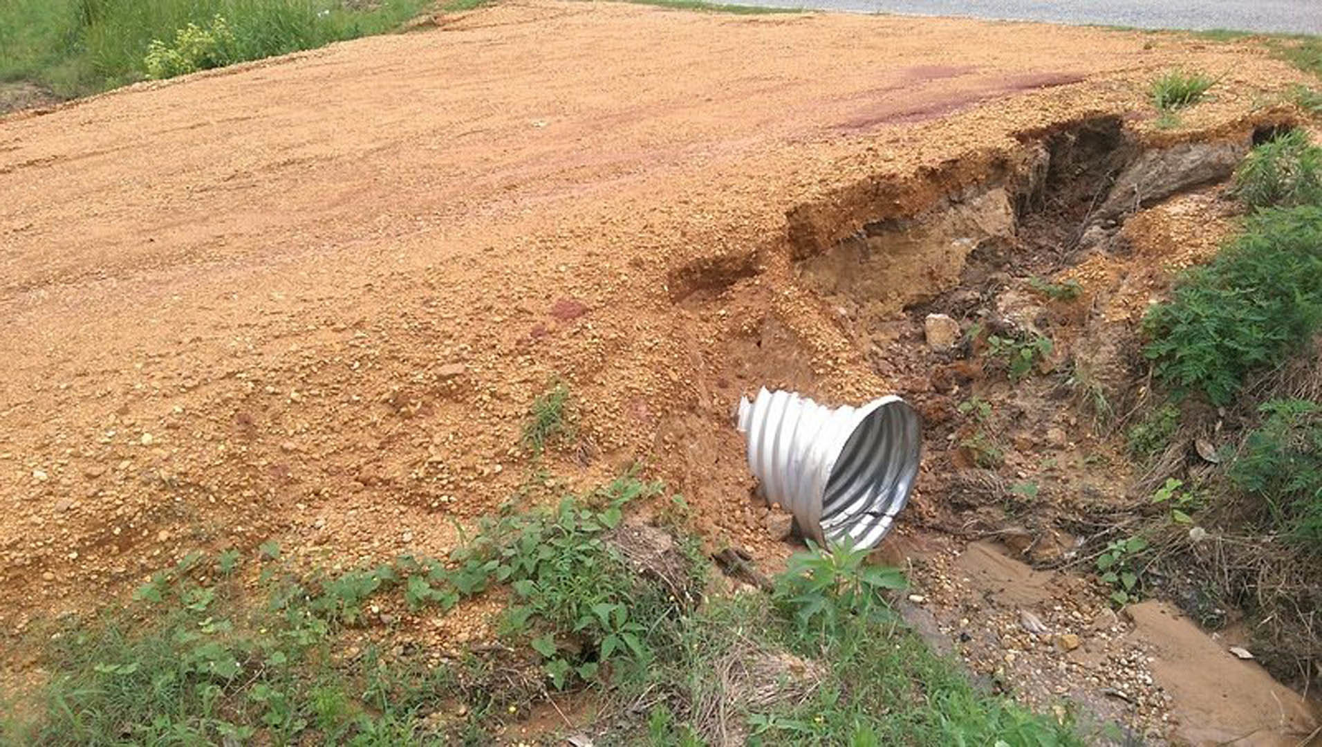 Metal pipe partially buried in soil and small rocks, surrounded by grass and plants in an outdoor ditch