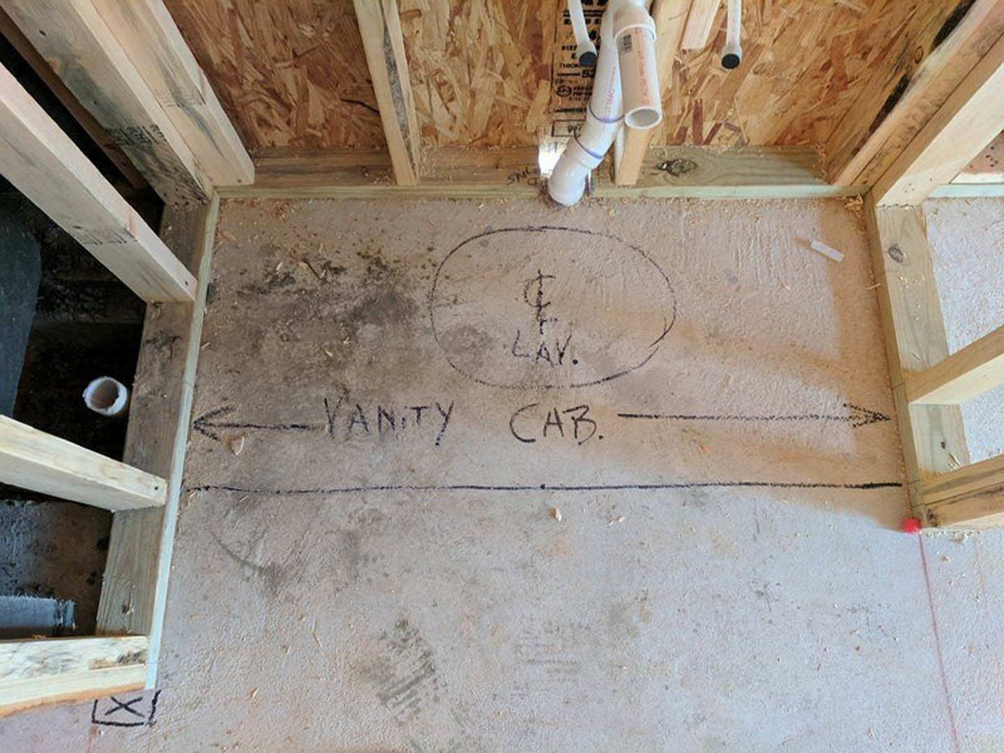 Bathroom under construction with exposed PVC pipes, unfinished concrete floor marked with handwritten notes, wood framing, and partially plastered walls