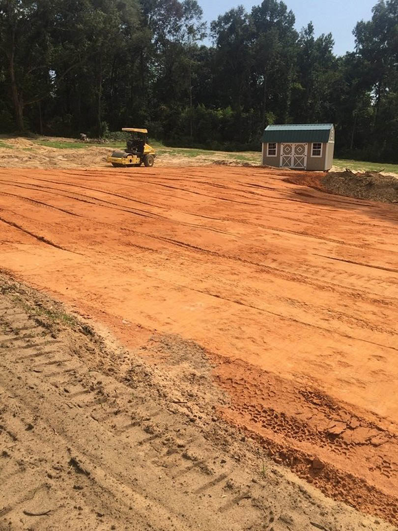 Partially built house with green roof, yellow construction vehicle with black cab parked on dirt ground, tire tracks visible, shed nearby, trees in background under open sky