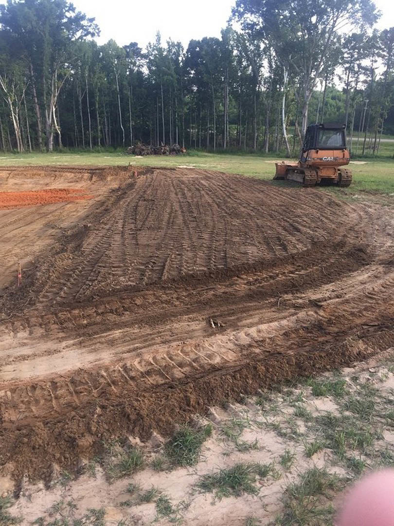 Tractor parked on freshly graded dirt field with tire tracks, surrounded by grass and scattered trees under a partly cloudy sky
