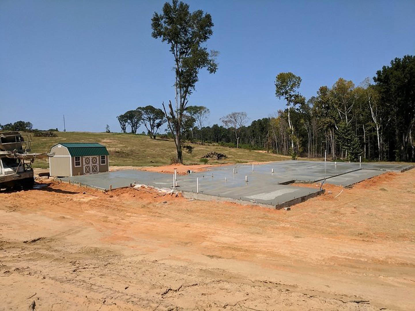 Concrete foundation and framing of a custom home under construction in an open field, with a small green-roofed shed, construction vehicle, and tall tree nearby