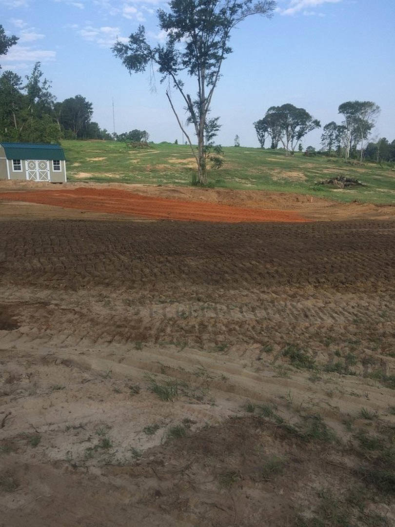 Single-story building with green metal roof sits beside a dirt field, surrounded by grass and scattered trees under a partly cloudy sky.