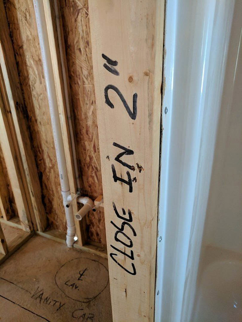Handwritten black text on a natural wood plank mounted indoors, with white wall and bathroom floor partially visible in background.