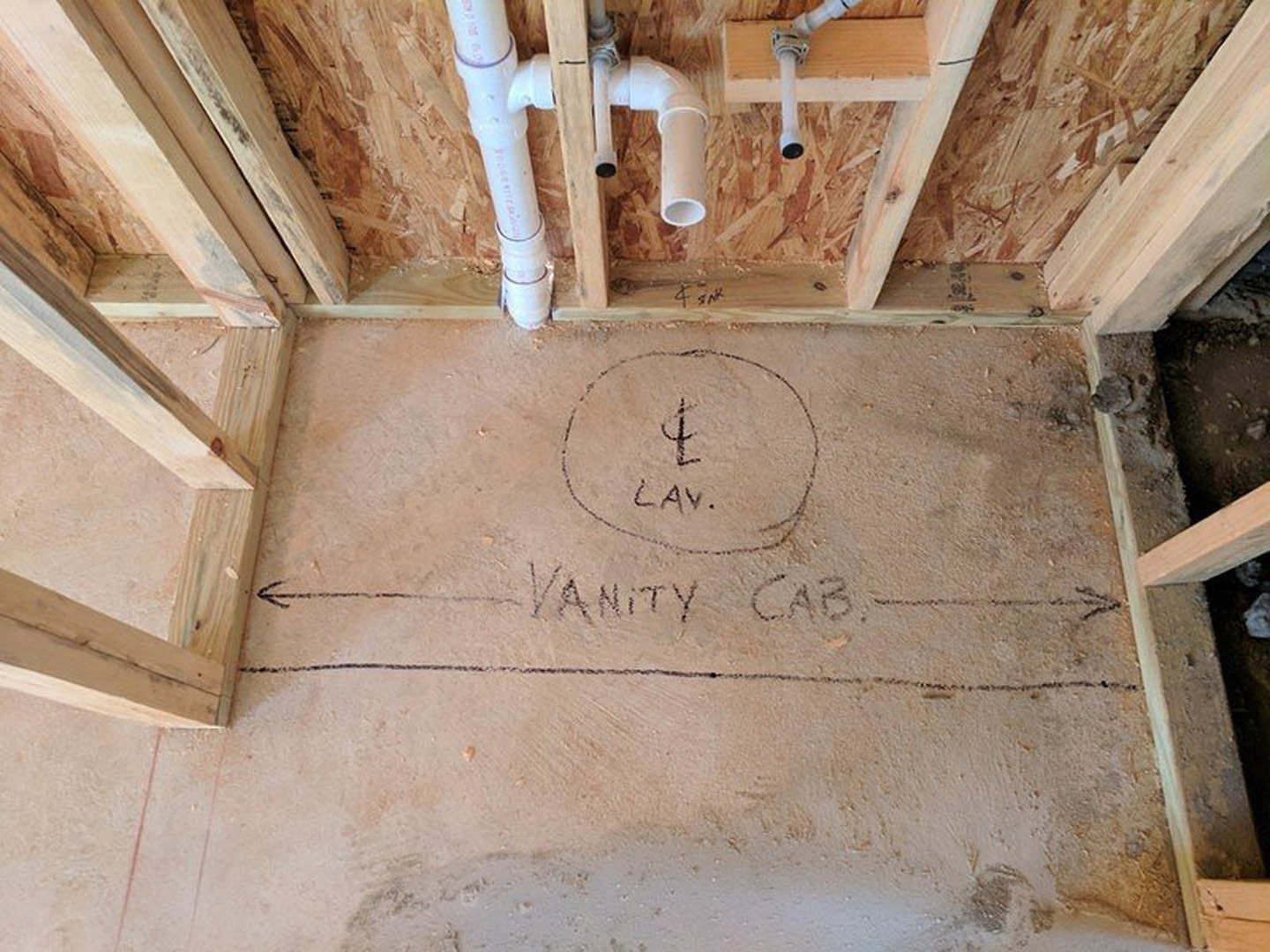 Beige bathroom under construction with exposed white PVC plumbing pipes attached to wood framing, unfinished plaster walls, and a sink area marked with handwritten notes on the