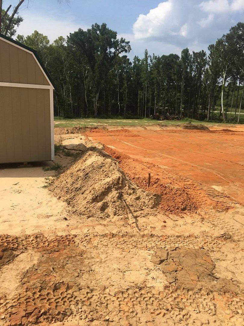 Large dirt pile with visible tire tracks beside a light-colored shed, open dirt field in foreground, wooden fence and cluster of trees in background under partly cloudy sky