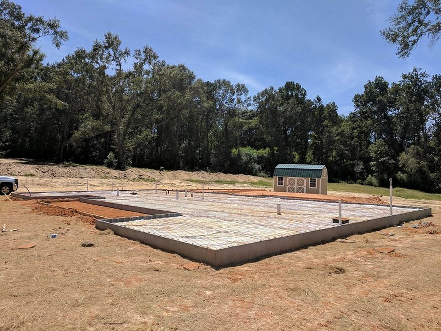 Partially built house with green roof, concrete slab foundation, construction truck parked nearby, surrounded by grassy field and scattered trees under cloudy sky