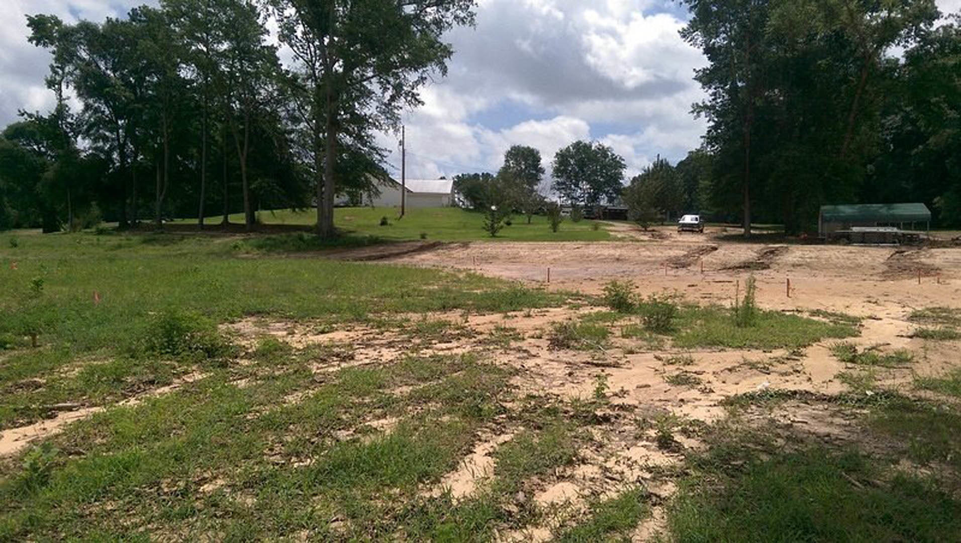White single-story building with flat roof set in a dirt field, surrounded by scattered trees and patches of grass under a cloudy sky