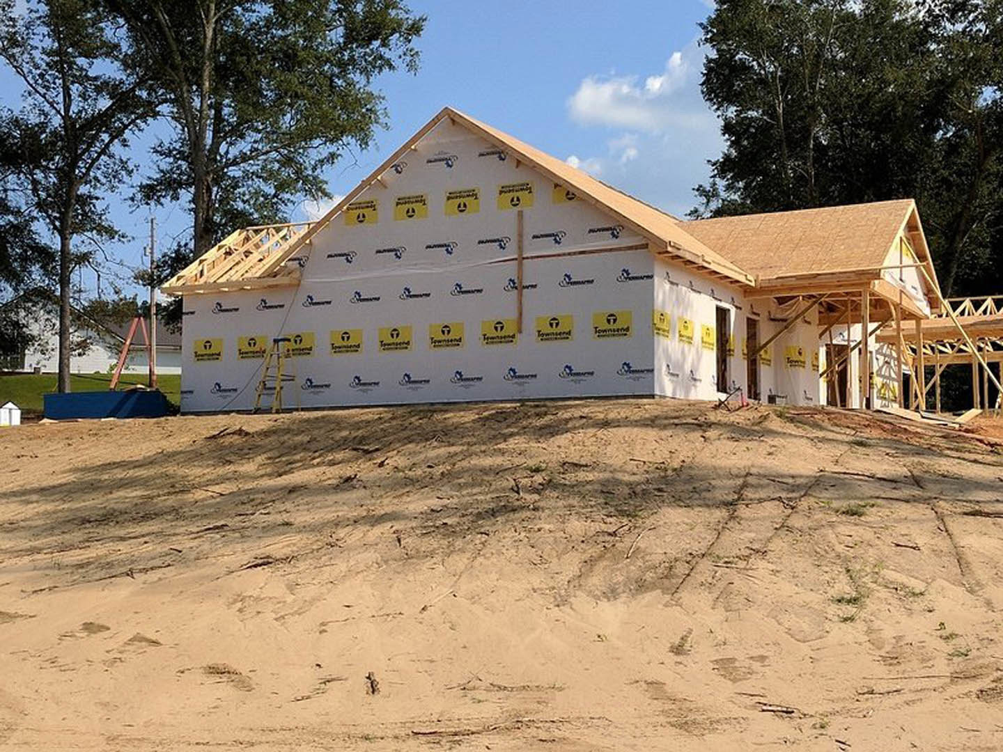 Partially built house with exposed framing, sandy ground, white exterior wall, blue tarp covering materials, yellow and black construction logos, leafy trees, and ladder leaning