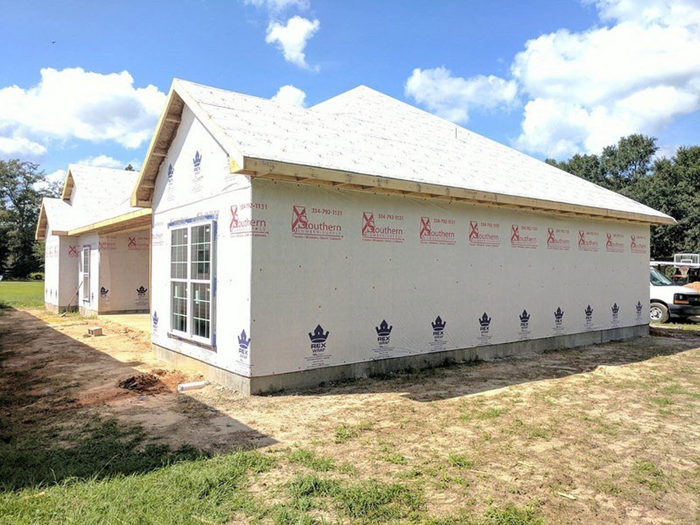 Two-story house under construction with exposed framing, shingled roof, and patch of green grass in front yard; large windows and white car parked nearby.