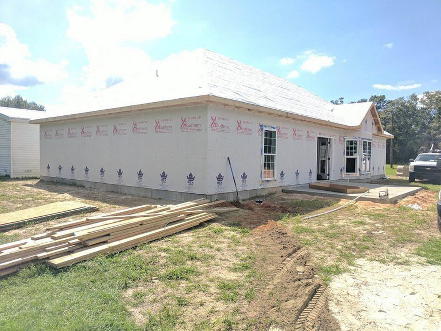 Wood-framed house under construction with exposed roof trusses, pile of lumber on grassy lot, white-framed window, cloudy sky overhead