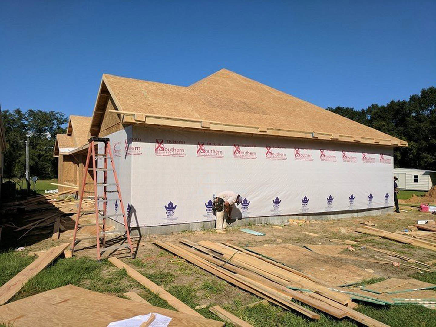 Man on ladder installing pipe beside partially built house with exposed framing, white shed with window in background, crown logo visible on wall.