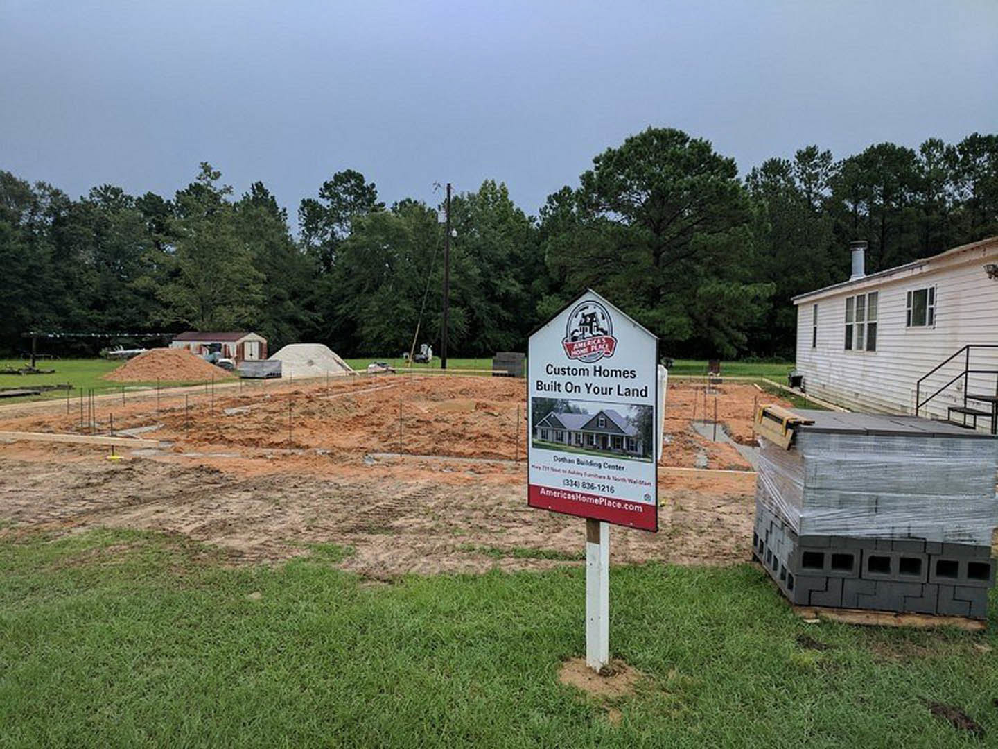 White modern home under construction with black metal staircase, stack of grey bricks, grassy lot, and a red and white logo sign in front of trees.