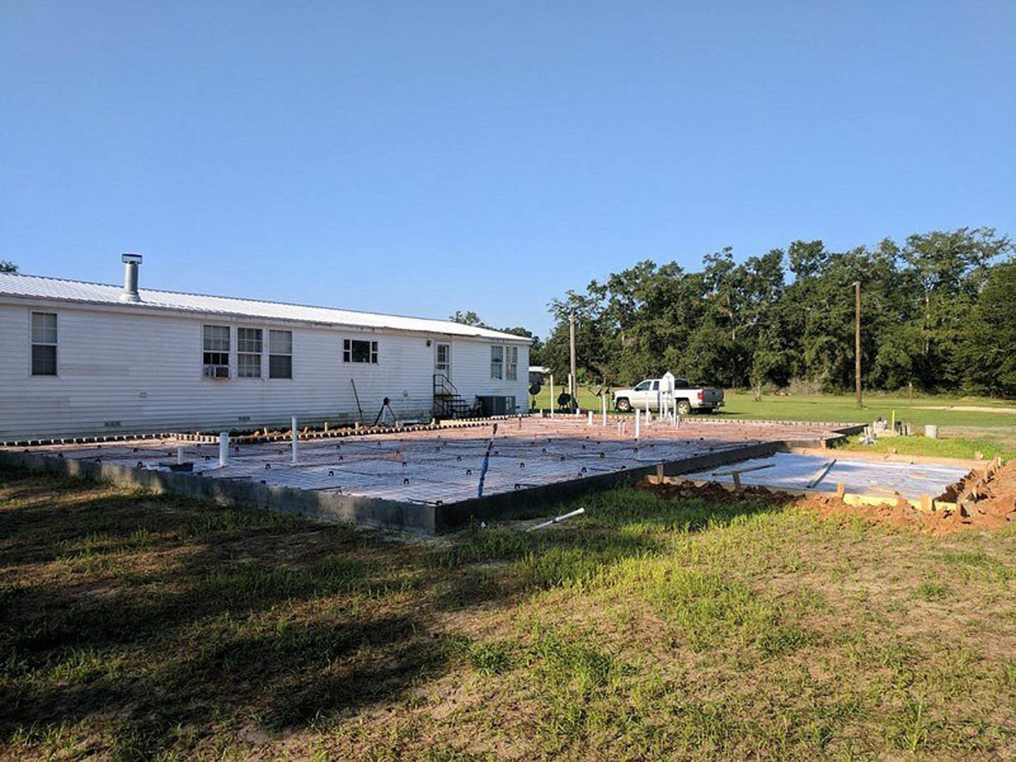 White house with multiple windows, concrete foundation slab with exposed metal bars, white truck parked nearby, grassy yard, group of trees in the background, blue sky with