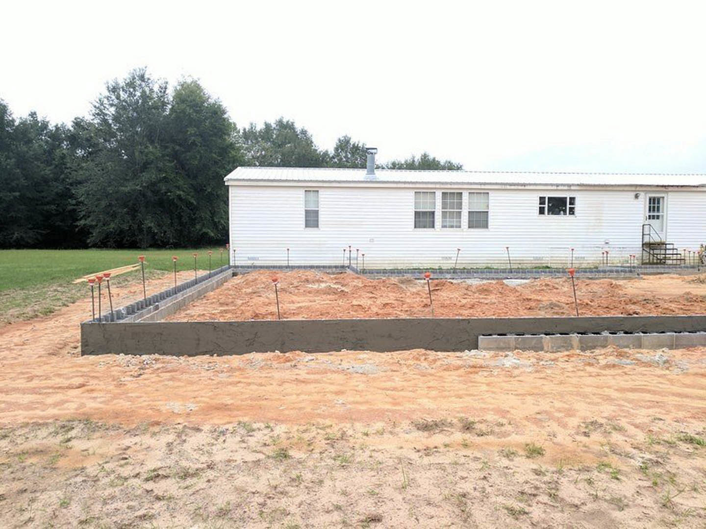White house with multiple windows, surrounded by leafy trees, sits on a dirt foundation with piles of earth and grass in the foreground.