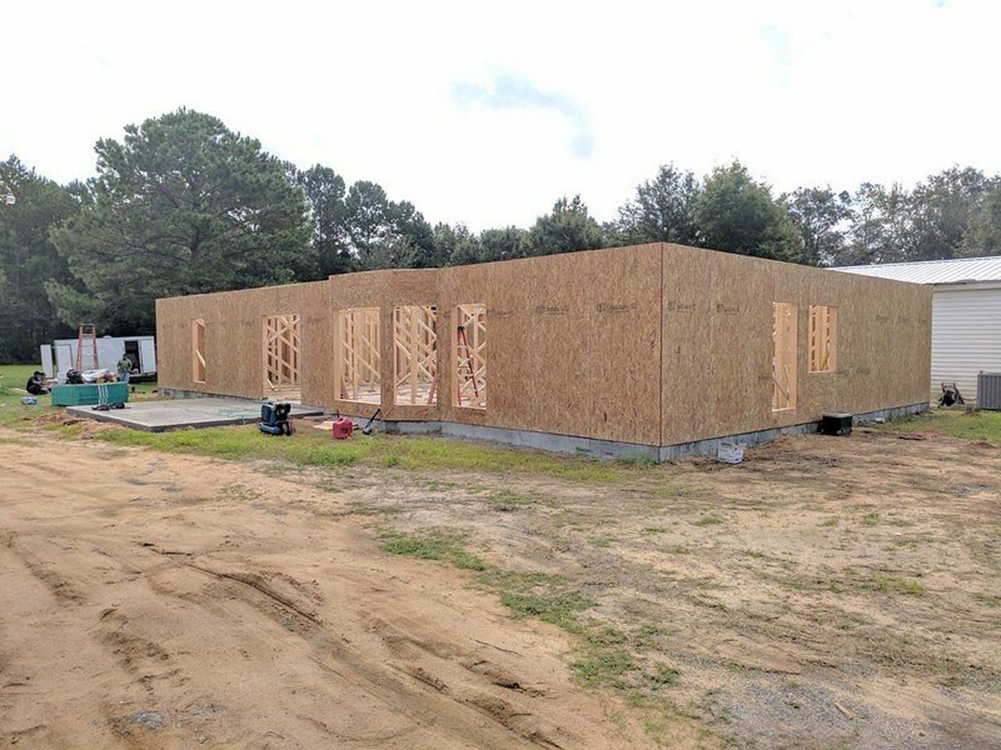 Two-story house under construction with exposed framing, dirt lot in foreground, garage opening visible, surrounded by trees and open sky