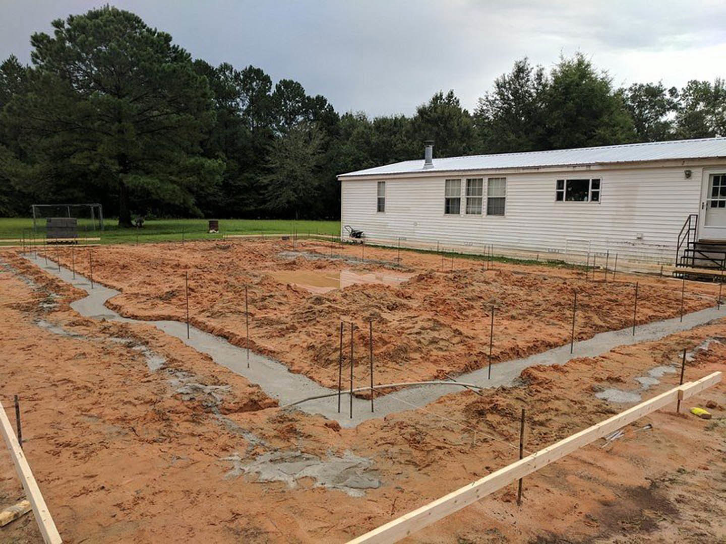 White house with multiple windows sits on exposed dirt foundation, surrounded by piles of soil and construction materials; grassy lot with scattered trees and cloudy sky in