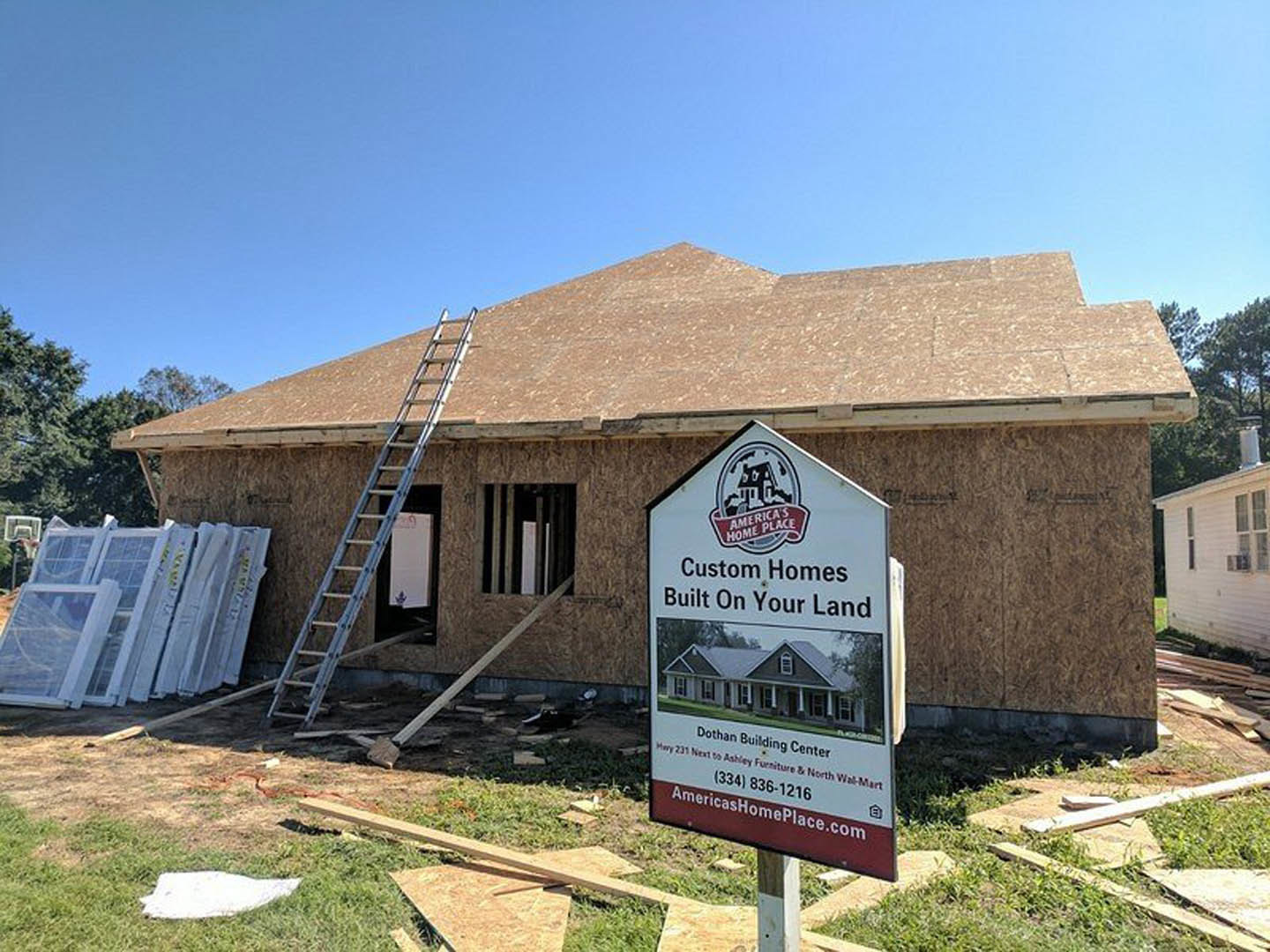 Framed house under construction with exposed wood beams, ladder leaning against roof, stack of windows nearby, construction sign displayed on exterior wall, surrounded by trees and