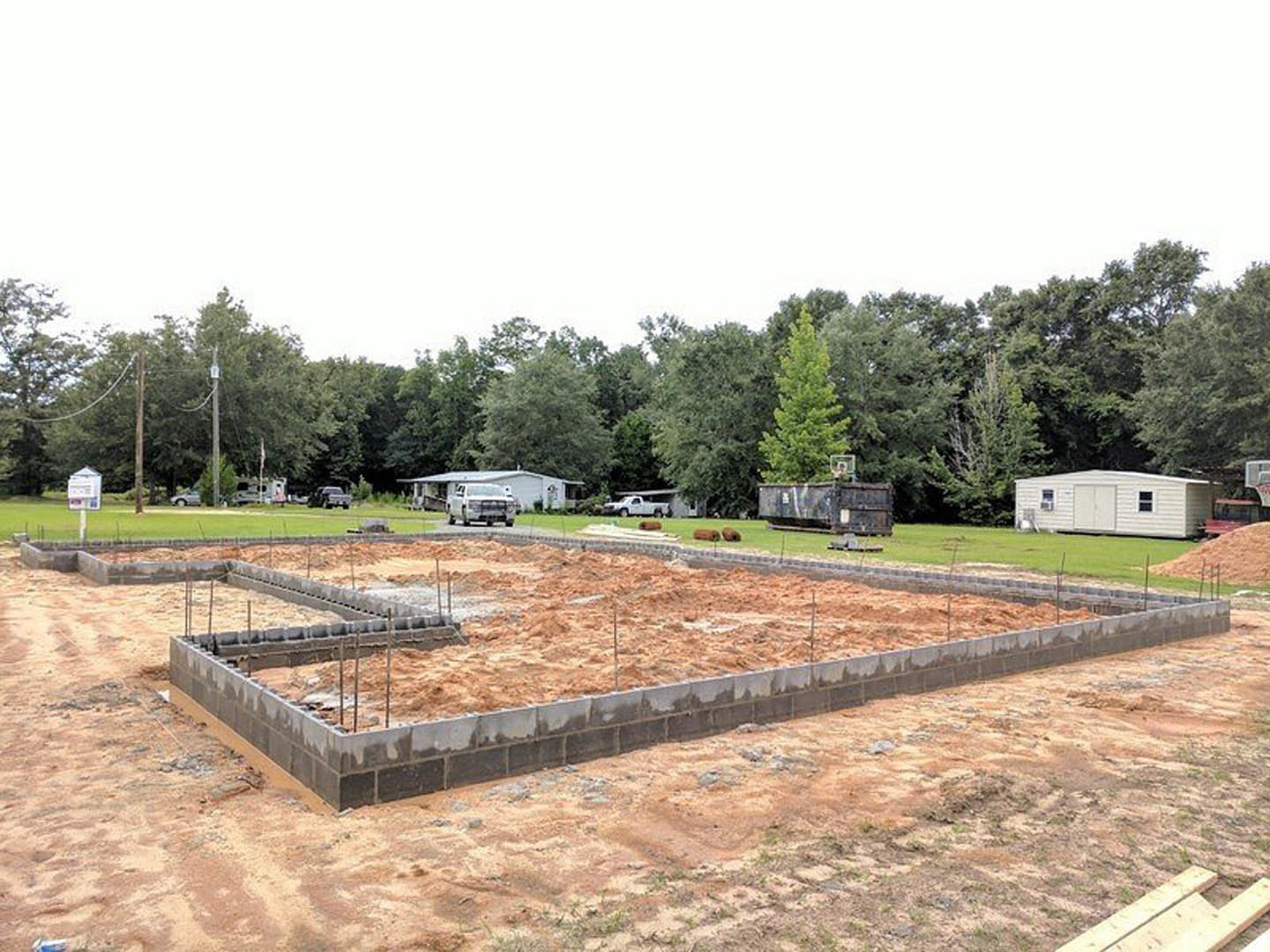 Concrete house foundation surrounded by dirt and grass, white shed with door in background, white van parked nearby, trees and building visible beyond construction site.