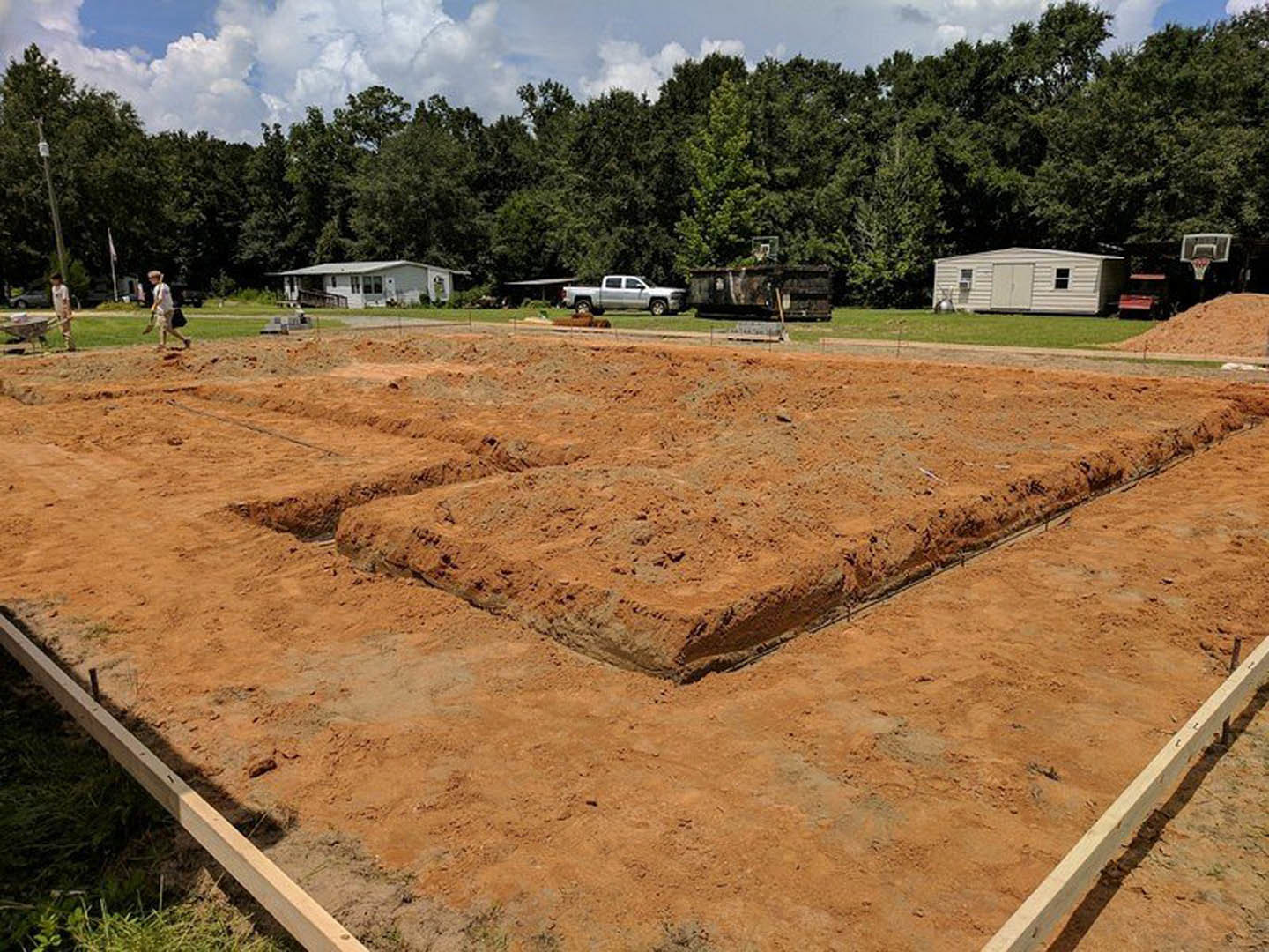 Modern white house with fenced yard, white shed, and white truck parked nearby; man standing in front, large dirt pile on ground, trees and cloudy sky in background