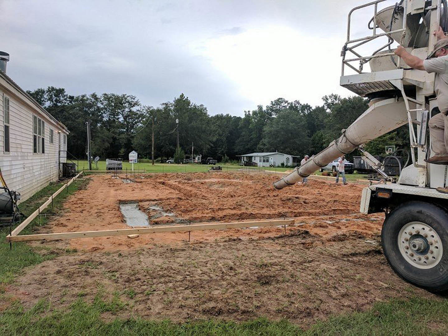 Cement truck pouring concrete onto dirt foundation area beside white house with mature trees and grassy yard