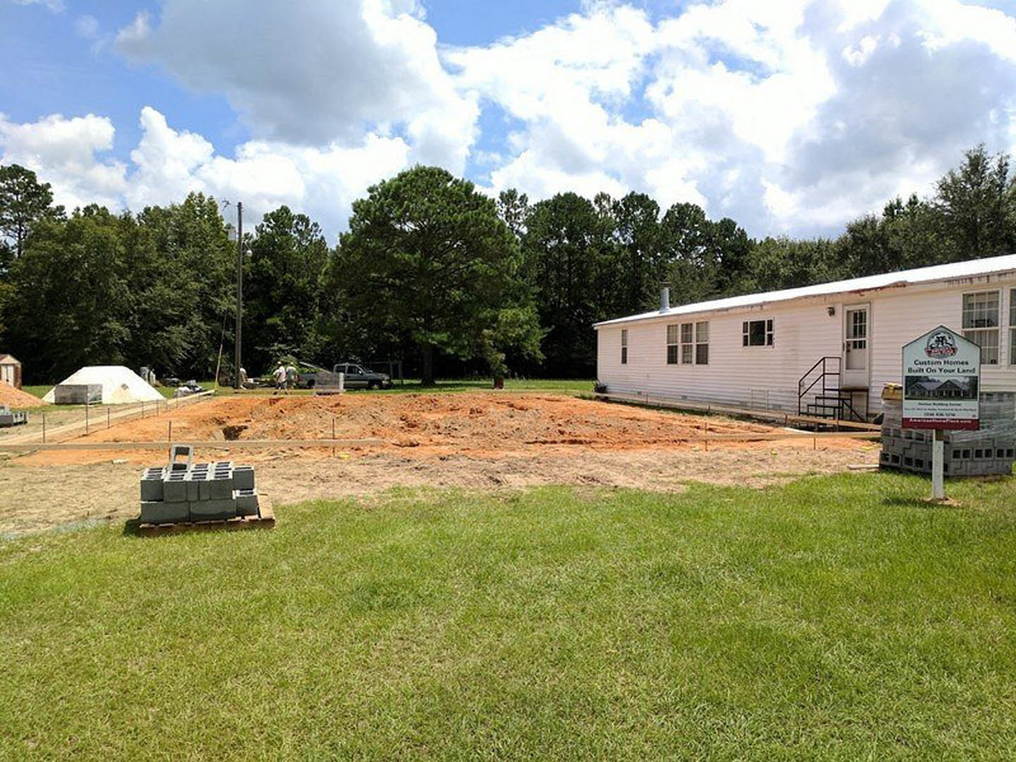 White two-story house with a prominent rear foundation, exterior staircase, and stacked grey bricks beside a grassy backyard under a partly cloudy sky.