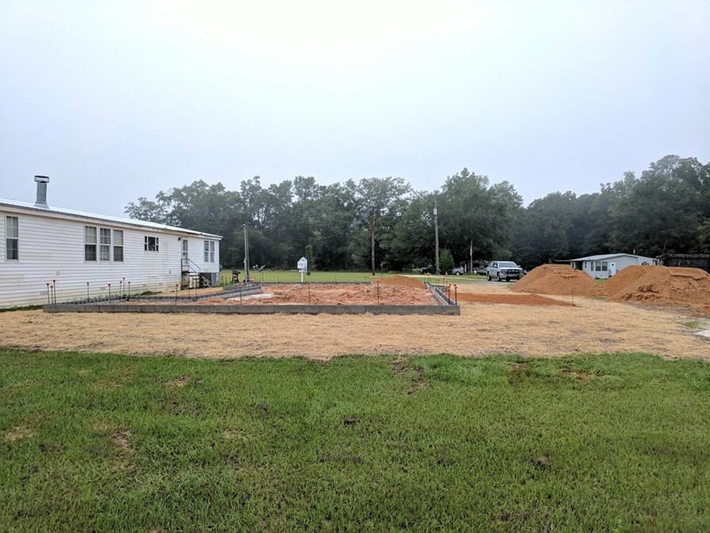 White house with red roof and chimney, surrounded by grass lawn and trees, dirt pile in front yard, blue sky overhead
