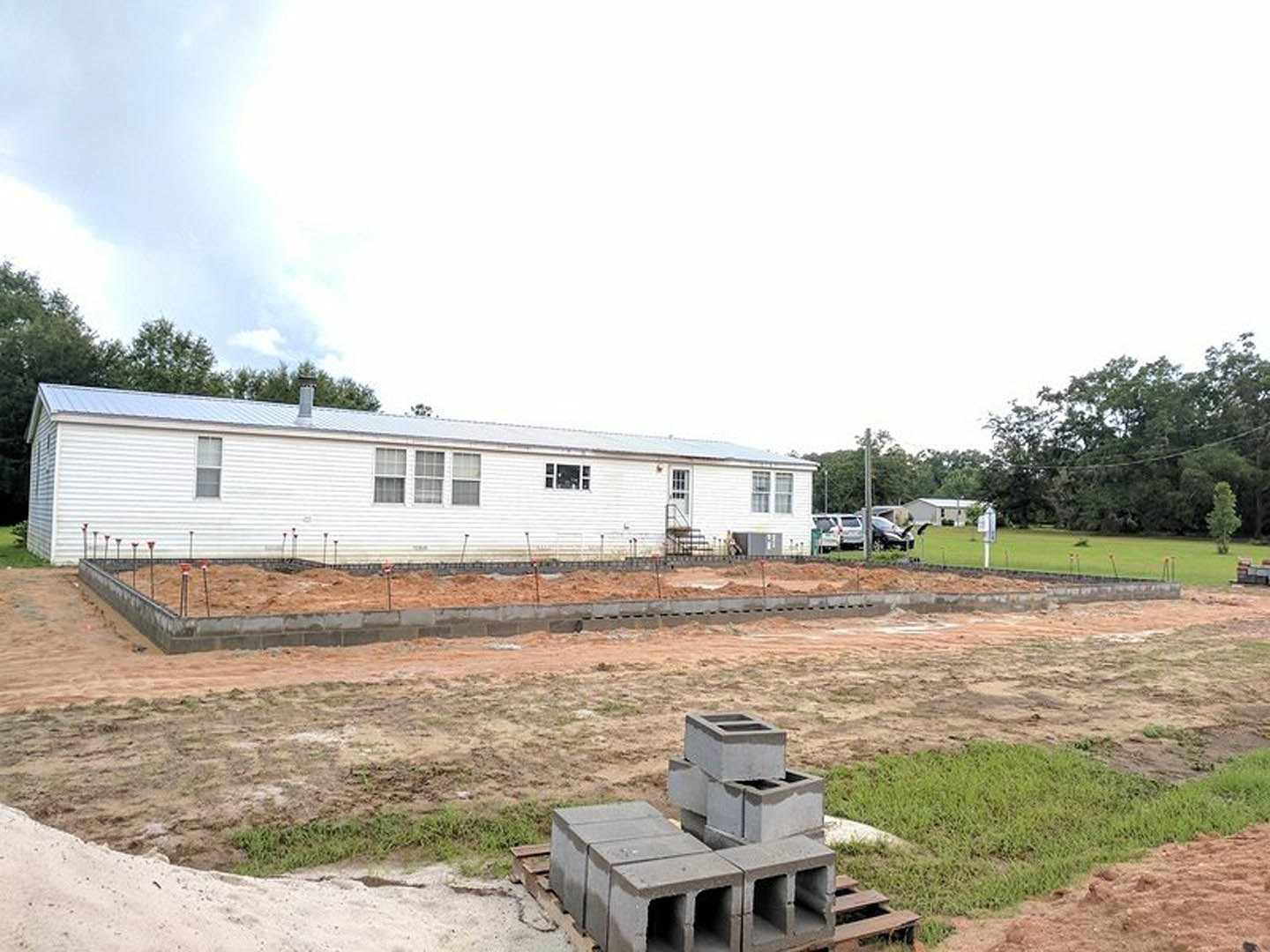 White house under construction with exposed concrete blocks, surrounded by grass and trees, person standing nearby, cloudy sky overhead