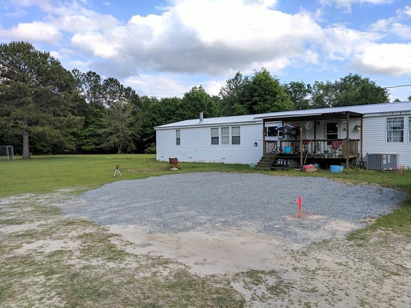 Gravel driveway leading to a house with a covered porch and stairs, grassy yard, trees, and partly cloudy sky