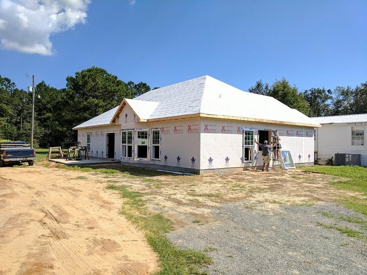 Framed custom home under construction with exposed roof trusses, ladder leaning against exterior wall, white-framed window installed, blue truck parked nearby, cloudy sky overhead