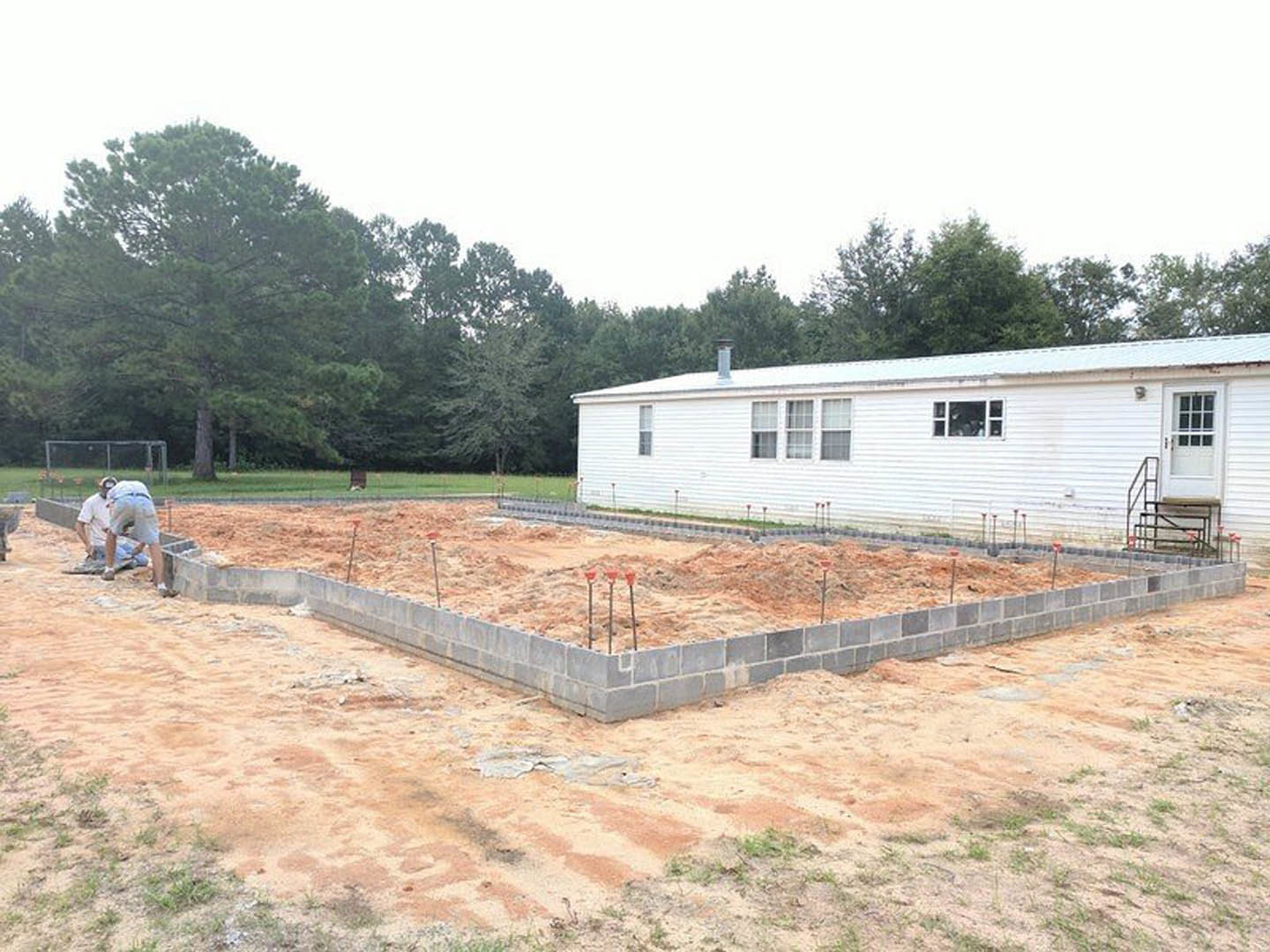 White house with multiple windows and glass-paneled door, surrounded by dirt and grass, foundation construction visible in foreground, two men working near the site, trees and