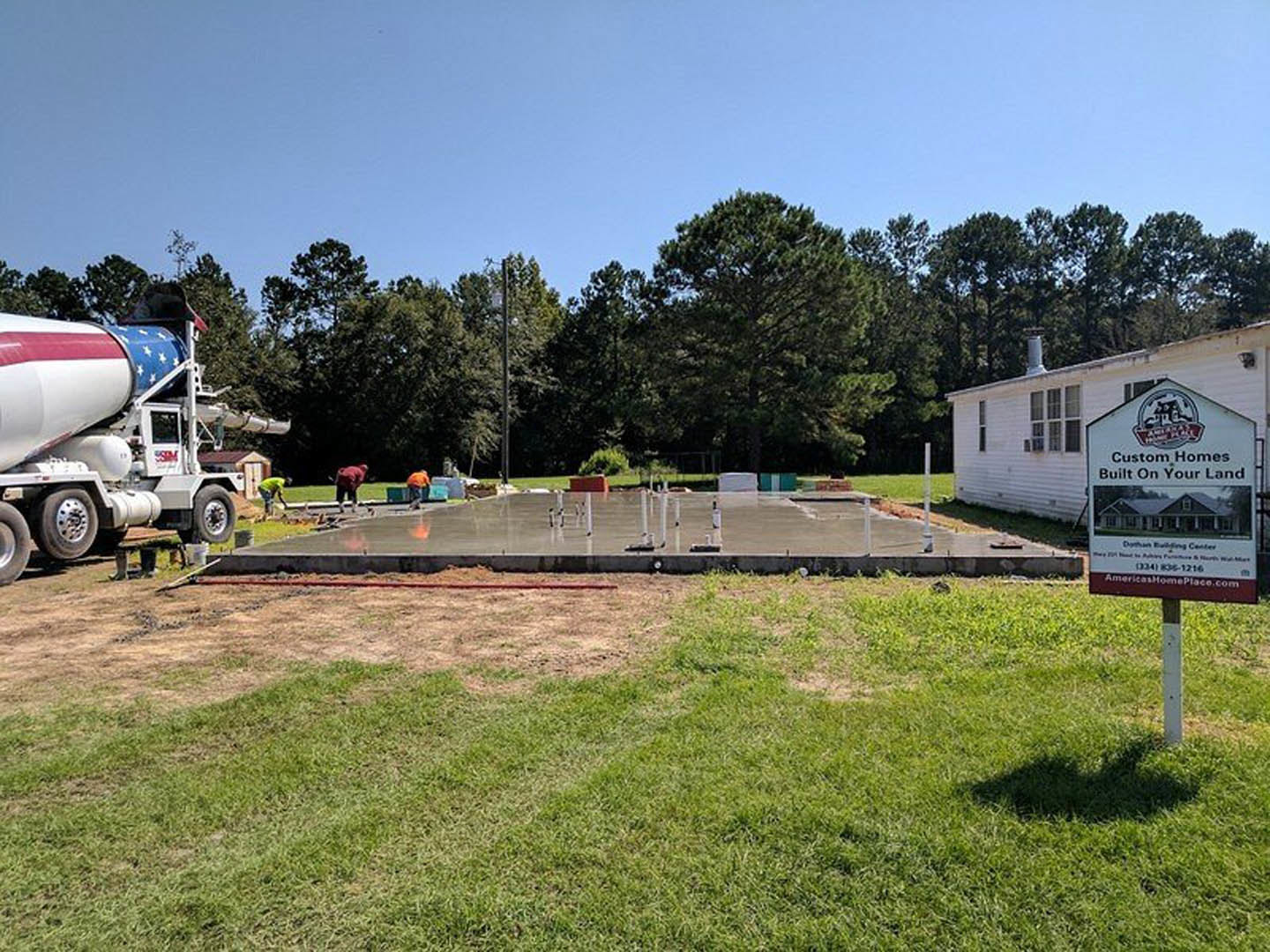 Concrete slab foundation and construction site beside a white house, grassy field, large tree, concrete mixer truck with flag, and signage displaying a house.