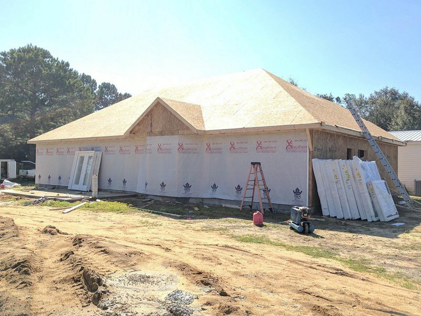 Partially built house with exposed framing, white door with glass panes, two ladders leaning against exterior, stack of white boards on ground, grassy lot with trees in background